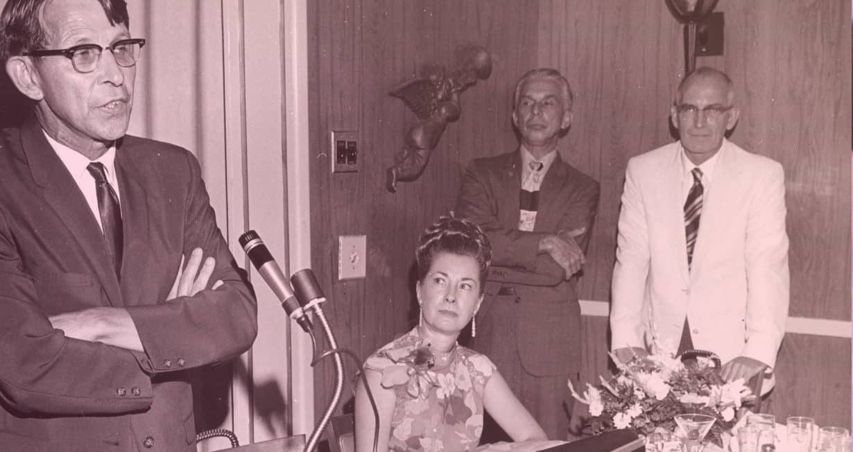 Photo Ancienne d'un homme les bras croisés parlant dans un micro. 2 hommes et une femme le regardent